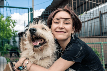 A female presenting person has her arms wrapped around a dog. They are sitting in front of the kennels at a dog shelter. The woman is smiling and the dog has it's mouth open. Both are facing the camera. 